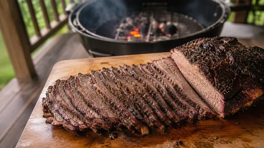 Sliced rested brisket on a wooden cutting board beside a grill with faint smoke in the background.
